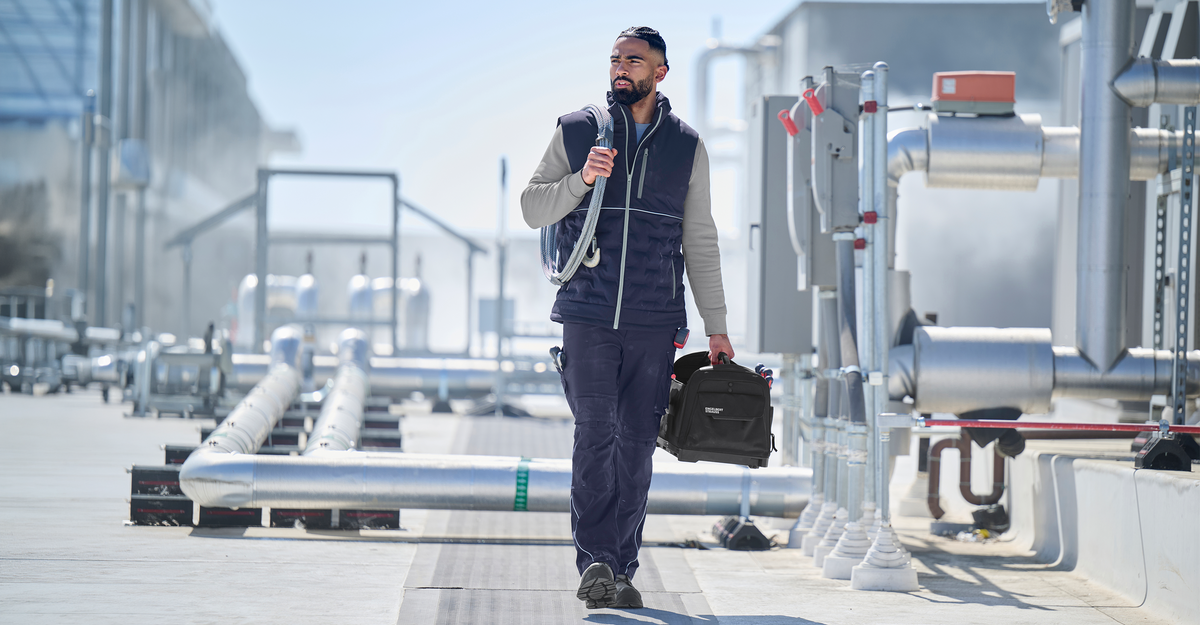 A man wearing navy workwear with platinum elements. He is walking on a rooftop with a tool box in his hand.