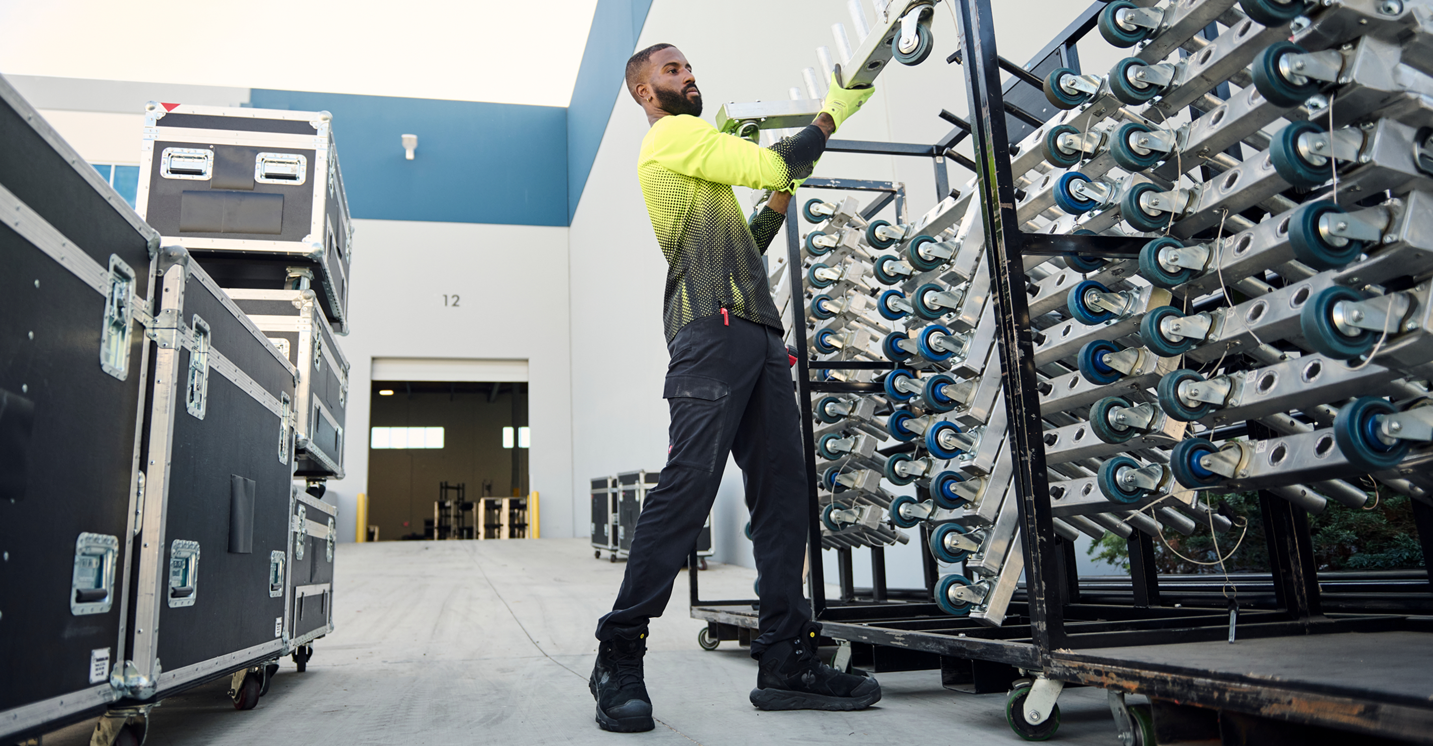 A man in a high-visibility neon yellow and black long-sleeve shirt, black pants, and black boots is handling a long metal pole on a wheeled cart filled with blue and white components in an industrial warehouse setting.
