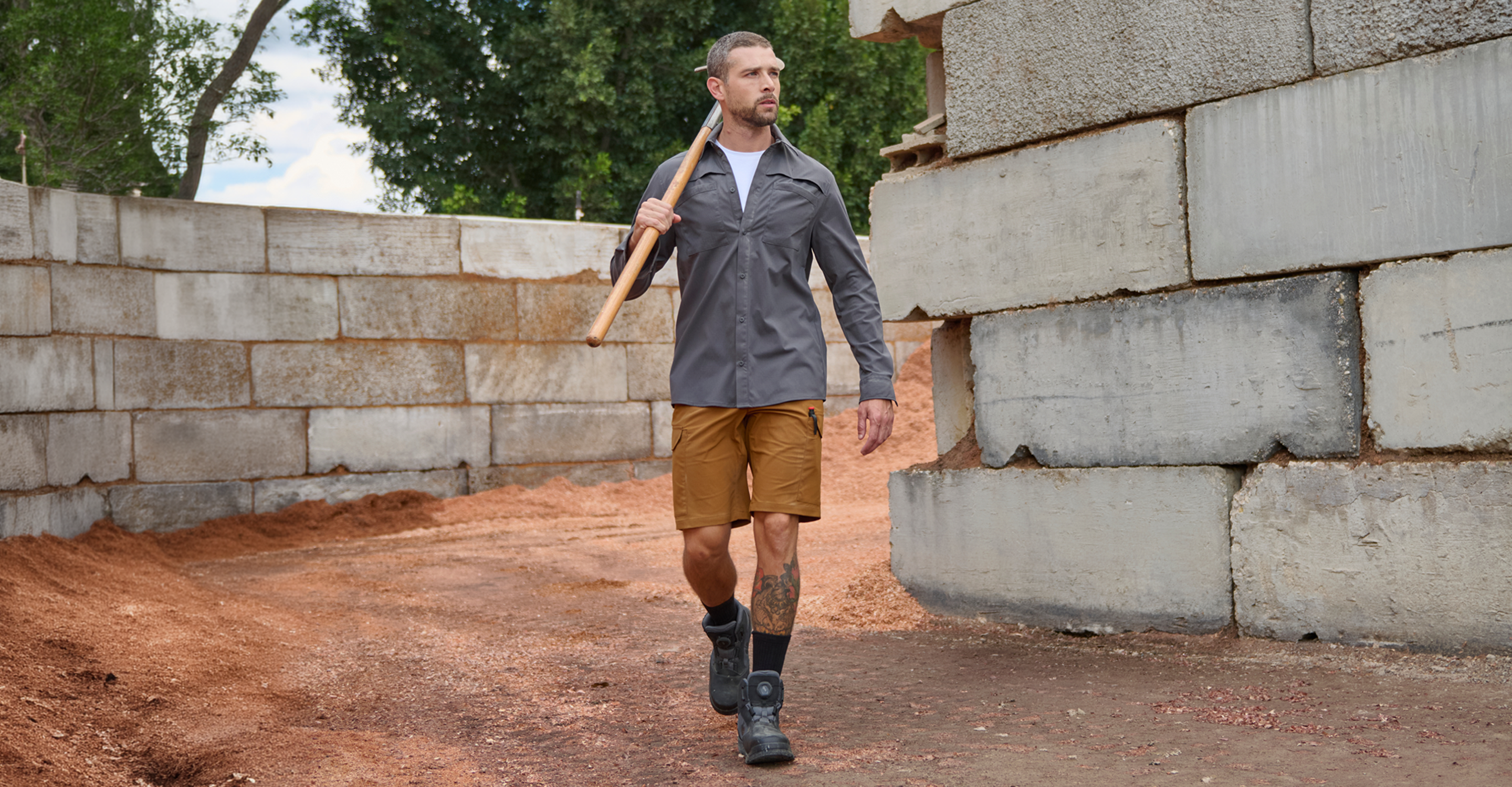 A man in a gray shirt, brown shorts, and black boots walks on a dirt path beside stone walls, carrying a shovel over his shoulder.