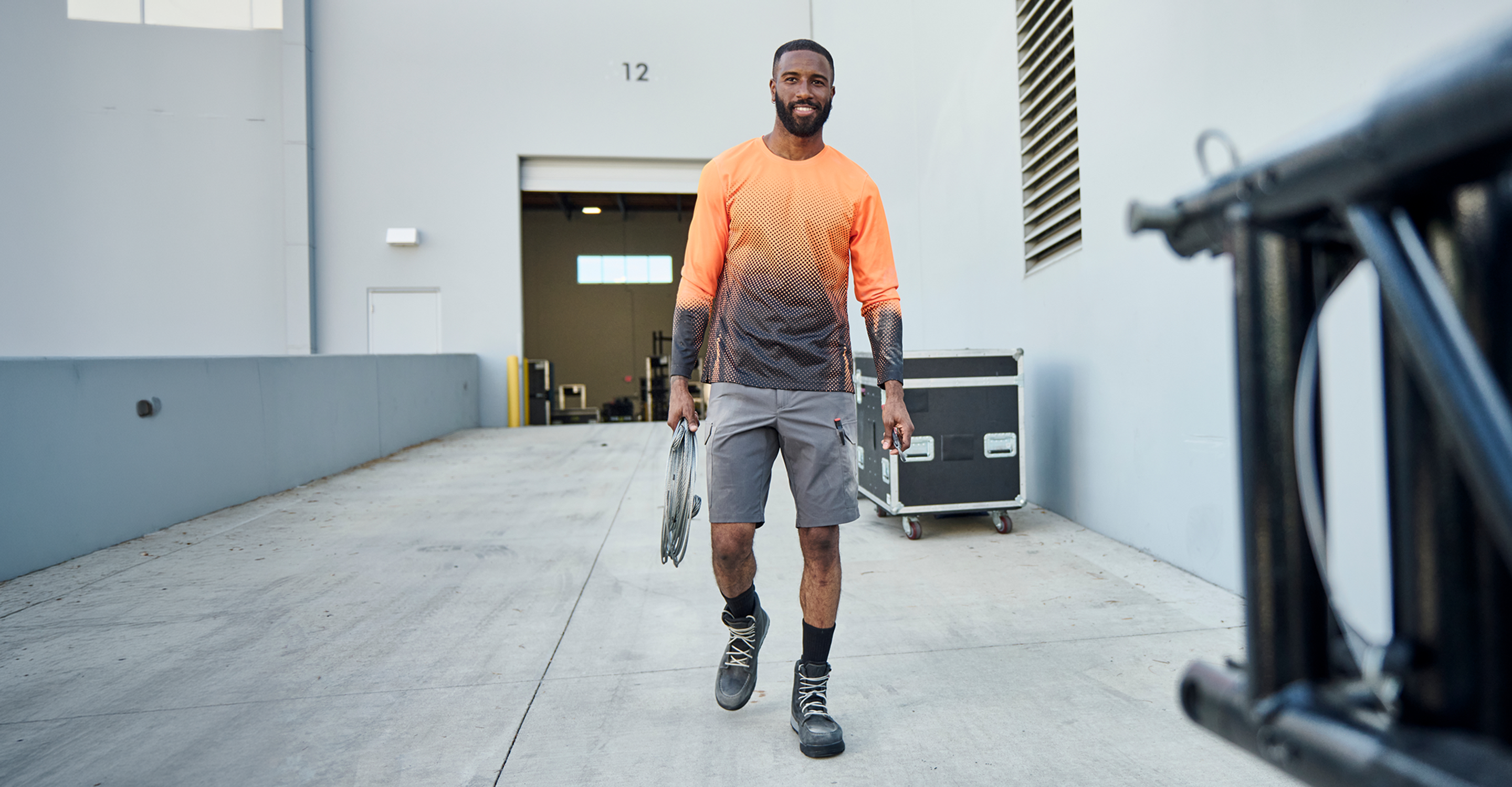 A man walking down a concrete industrial corridor wearing a long-sleeve athletic shirt with a bright orange-to-dark gray gradient, gray shorts, and black athletic shoes, while carrying a disc golf disc.