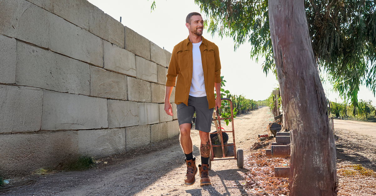 A man in a brown jacket, white shirt, shorts, and boots walks down a sunny dirt path beside a stone wall, with trees and a wheelbarrow in the background.