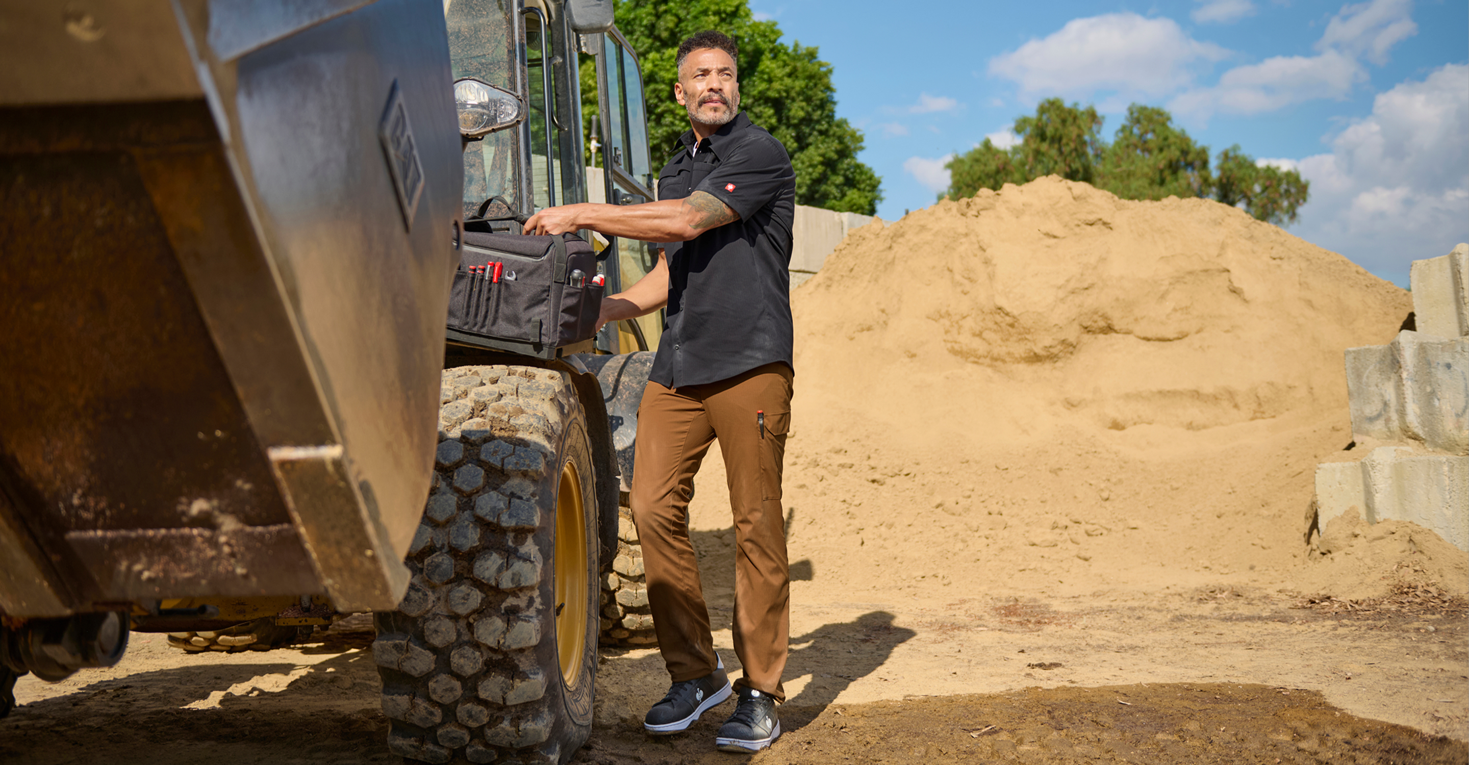 A man in work clothes stands next to heavy construction equipment on a sandy work site, holding a tool bag. There are large piles of sand and concrete blocks in the background under a blue sky.