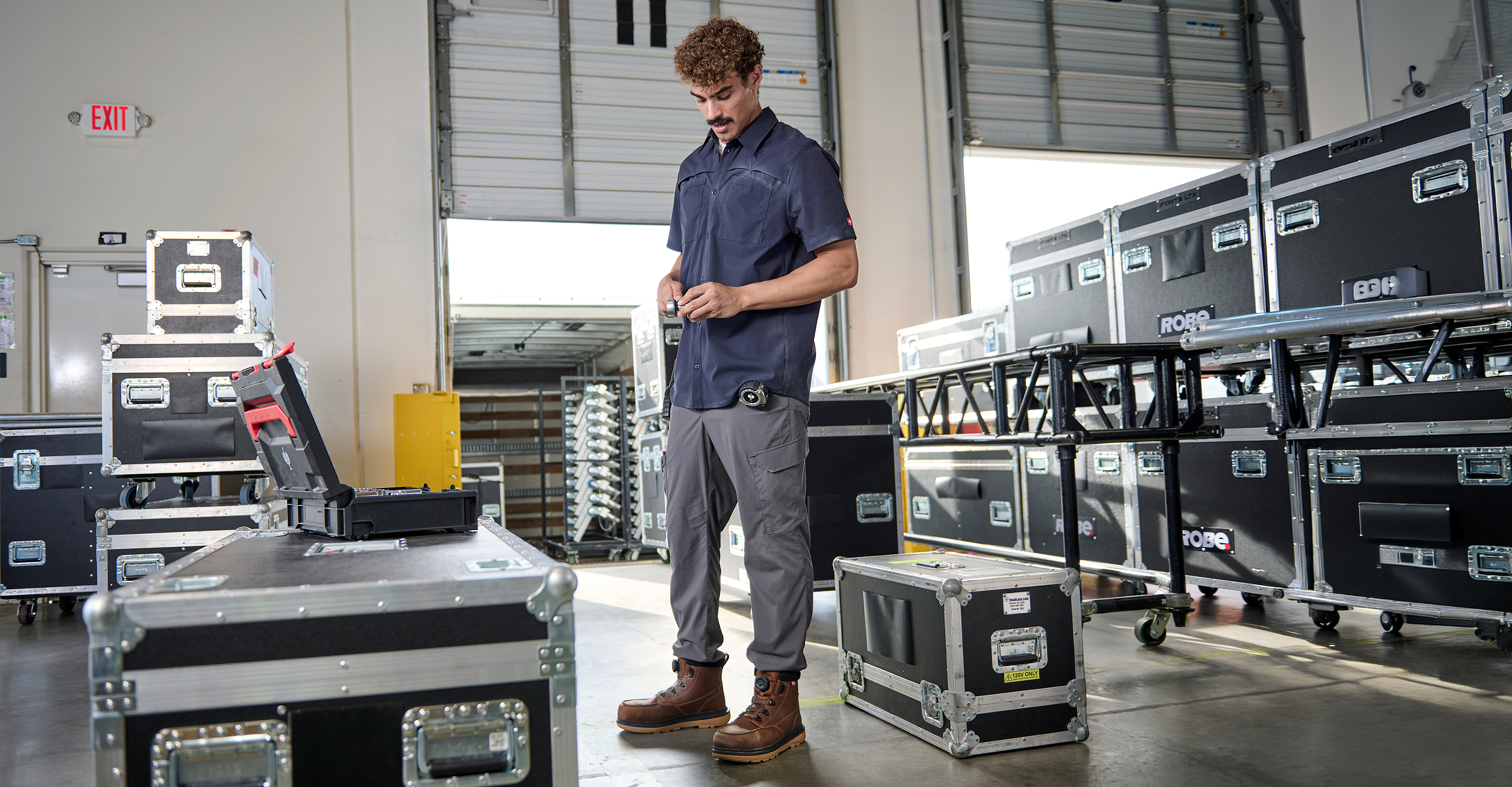 A man in a warehouse stands next to a black and gray audio equipment travel case, with stacks of similar cases in the background.