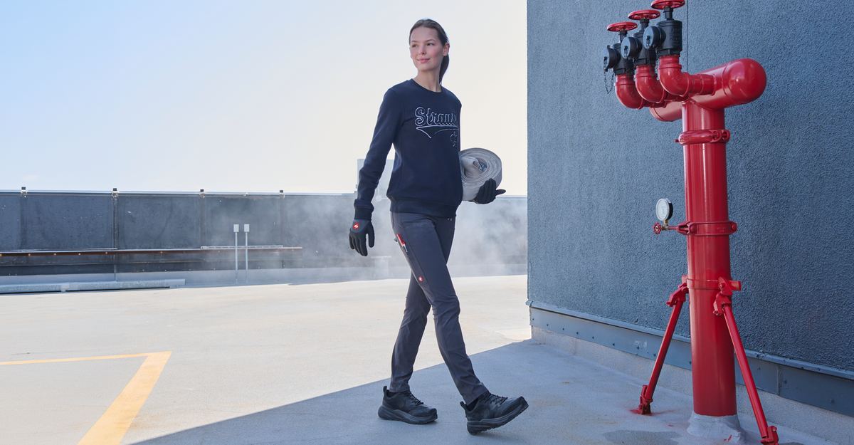 A woman in work attire walks on a rooftop holding a coiled hose, with red fire pipes and a gray wall nearby. Steam rises in the background.