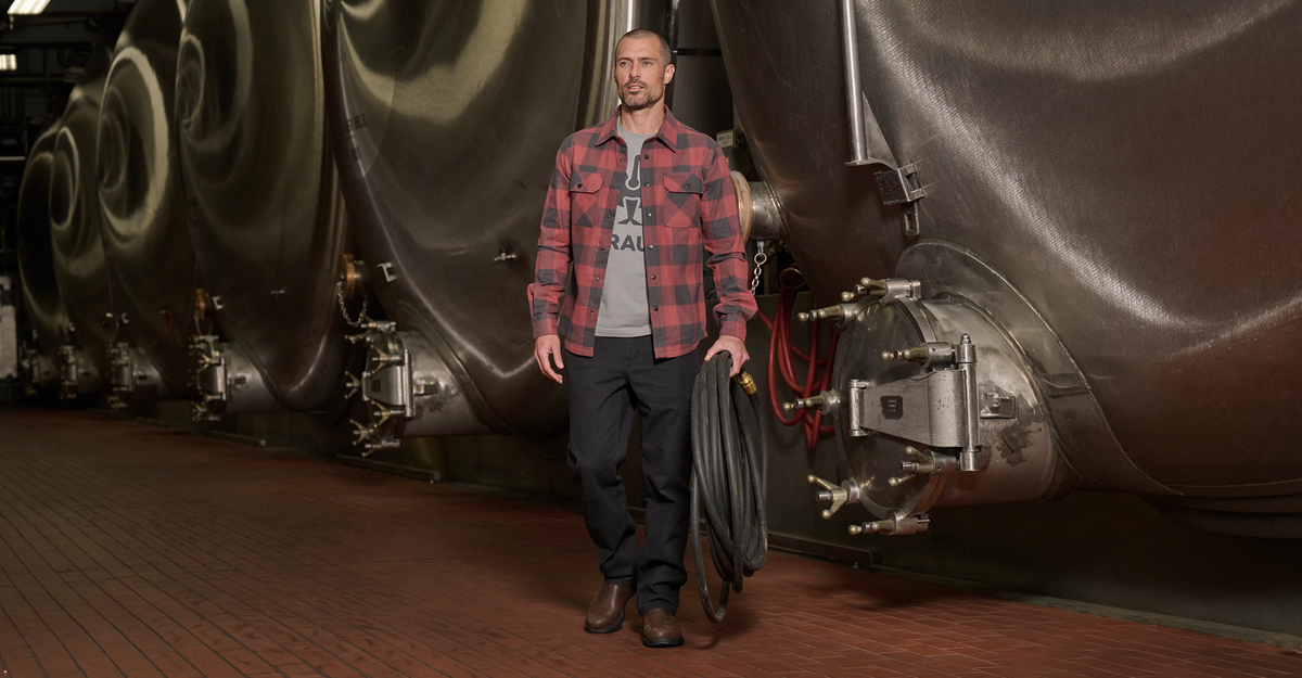 A man in a plaid shirt, gray t-shirt, and boots walks inside an industrial brewery, holding a large coiled hose. Stainless steel fermentation tanks line the background.