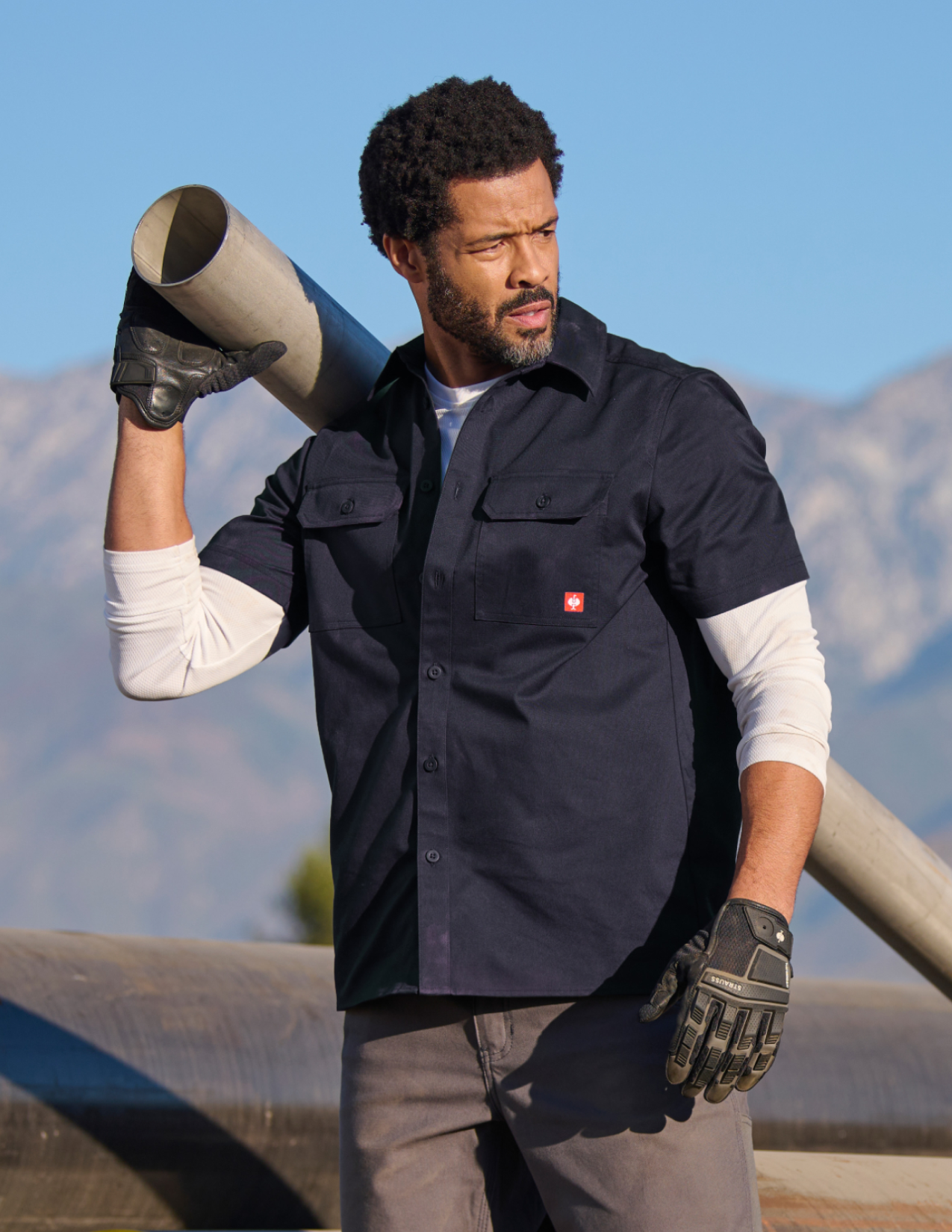 Man in navy work shirt and gloves holding a cylindrical object with mountains in the background