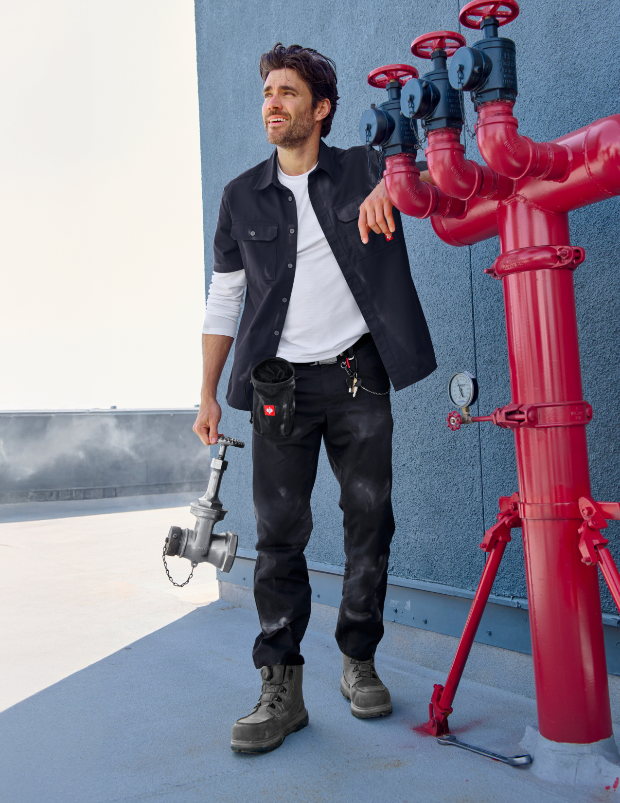 Man holding a wrench next to red pipes against a blue wall wearing a navy work shirt
