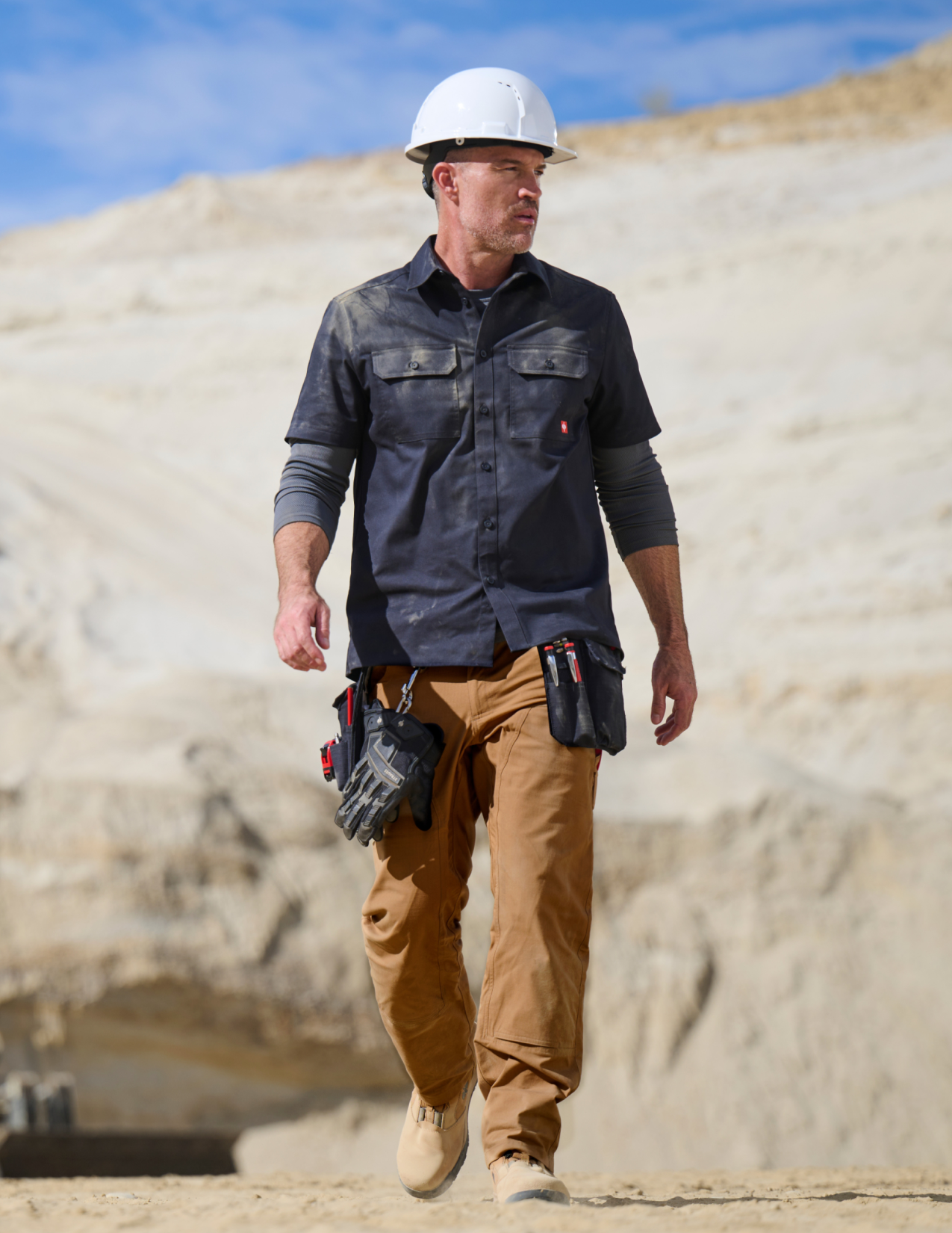Man wearing a navy work shirt with a hard hat walking on a construction site.
