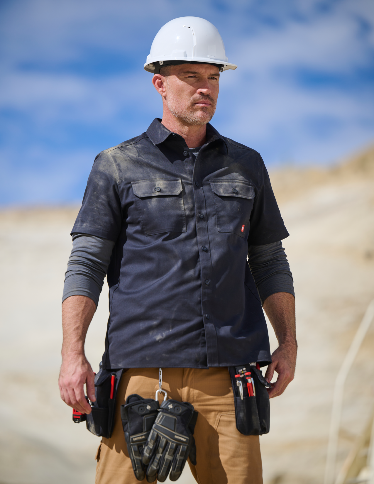 Man wearing a hard hat and navy work shirt with tools, standing outdoors.