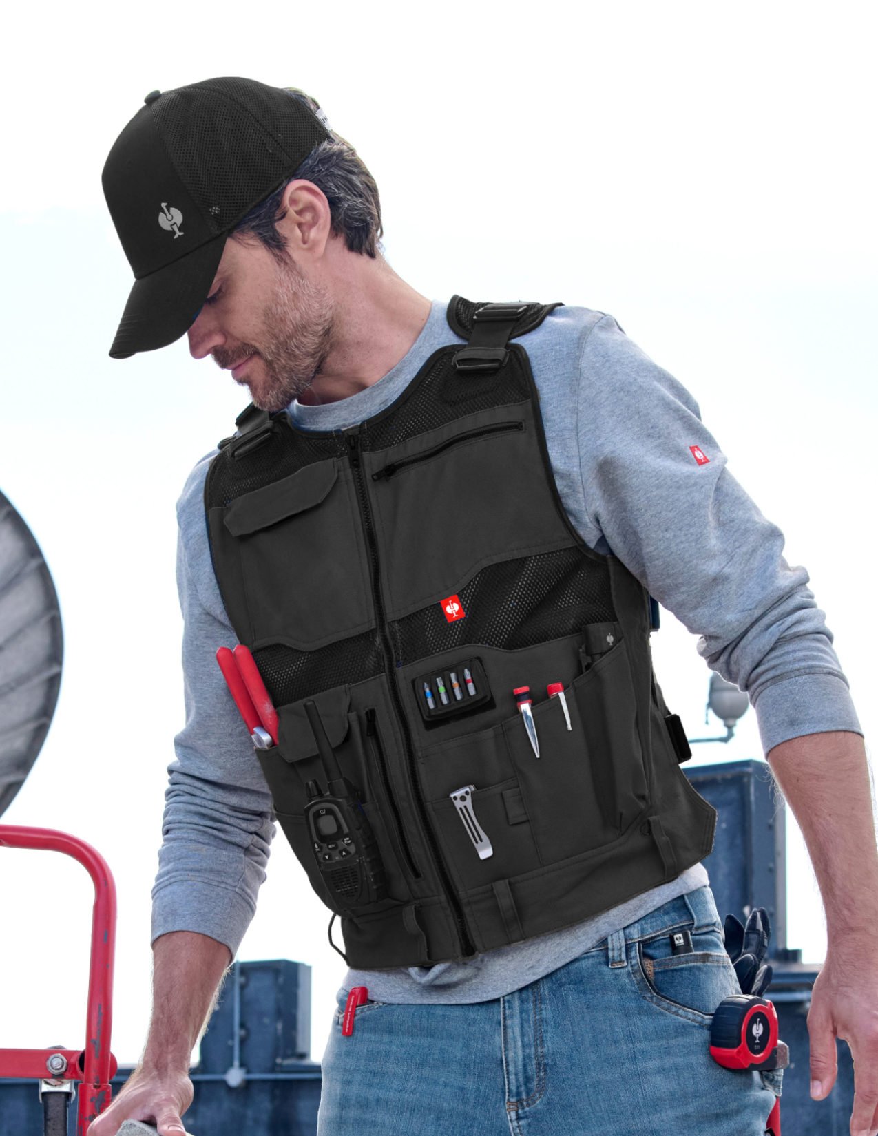 Man wearing a black tool vest with various pouches and tools on a white background