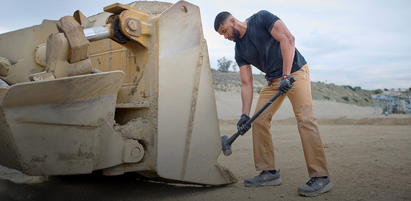 Man using a sledgehammer on a large piece of construction equipment in chinos.