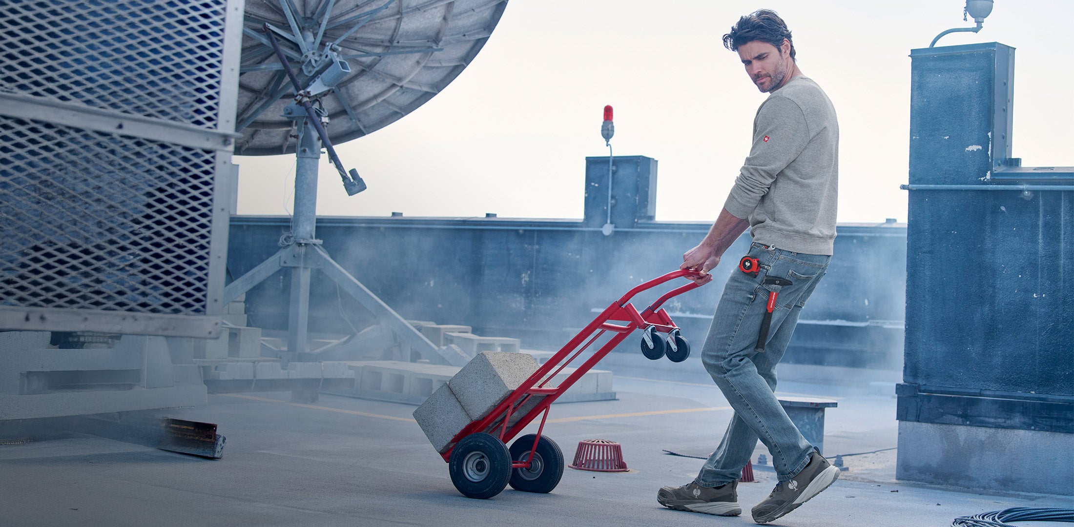 Person using a red hand truck on a rooftop with satellite equipment in the background in Power Denim.