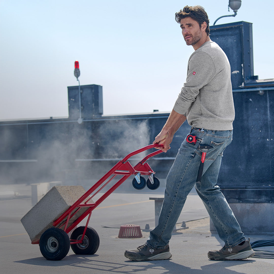 Man pushing a red hand truck on a rooftop with a clear sky in power denim. 